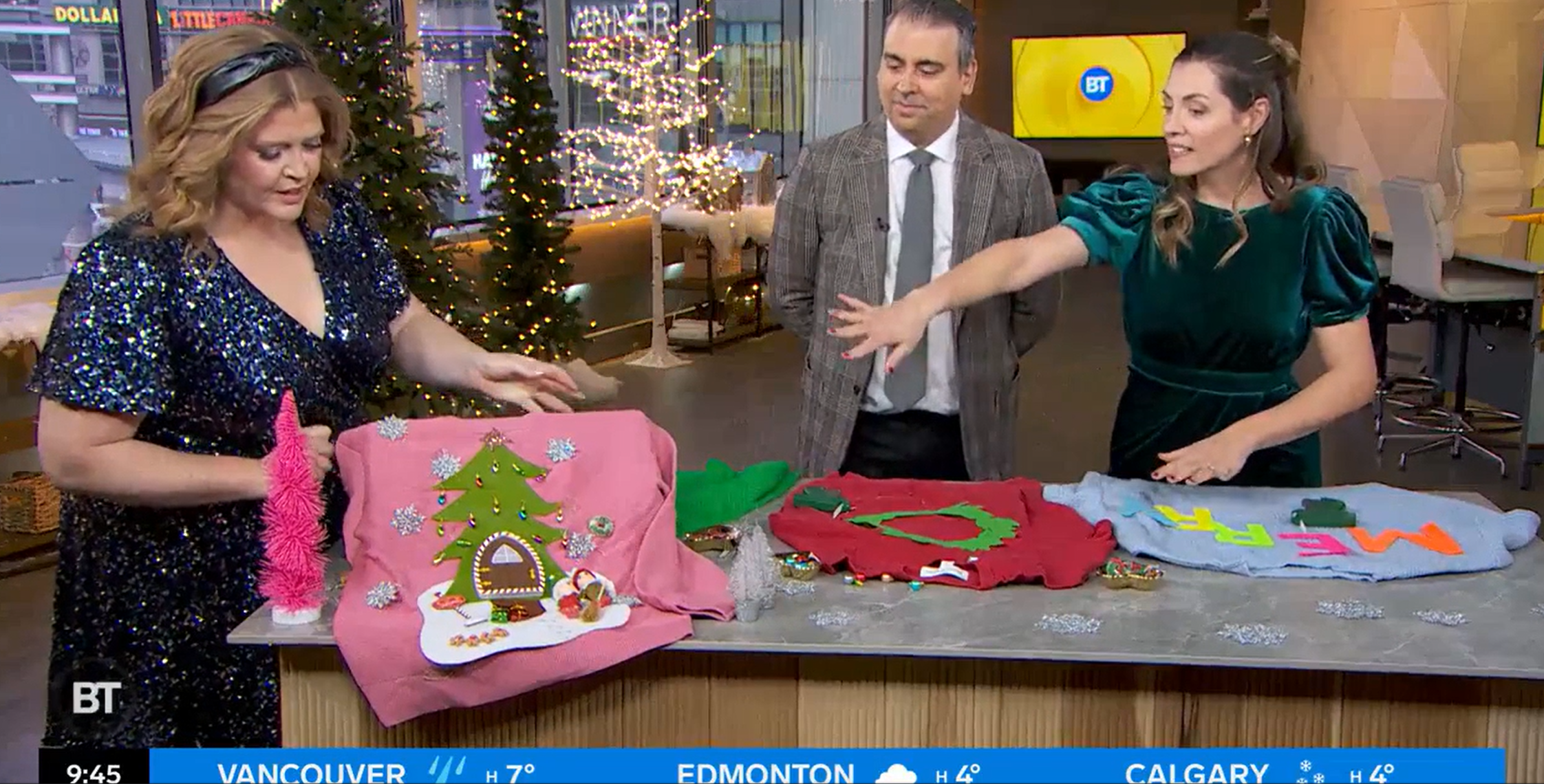 Three people stand around a table displaying festive sweaters with colorful decorations. The person on the left holds a pink sweater with a decorated Christmas tree, while the persons in the center and right observe and gesture about the sweaters. The setting appears festive.