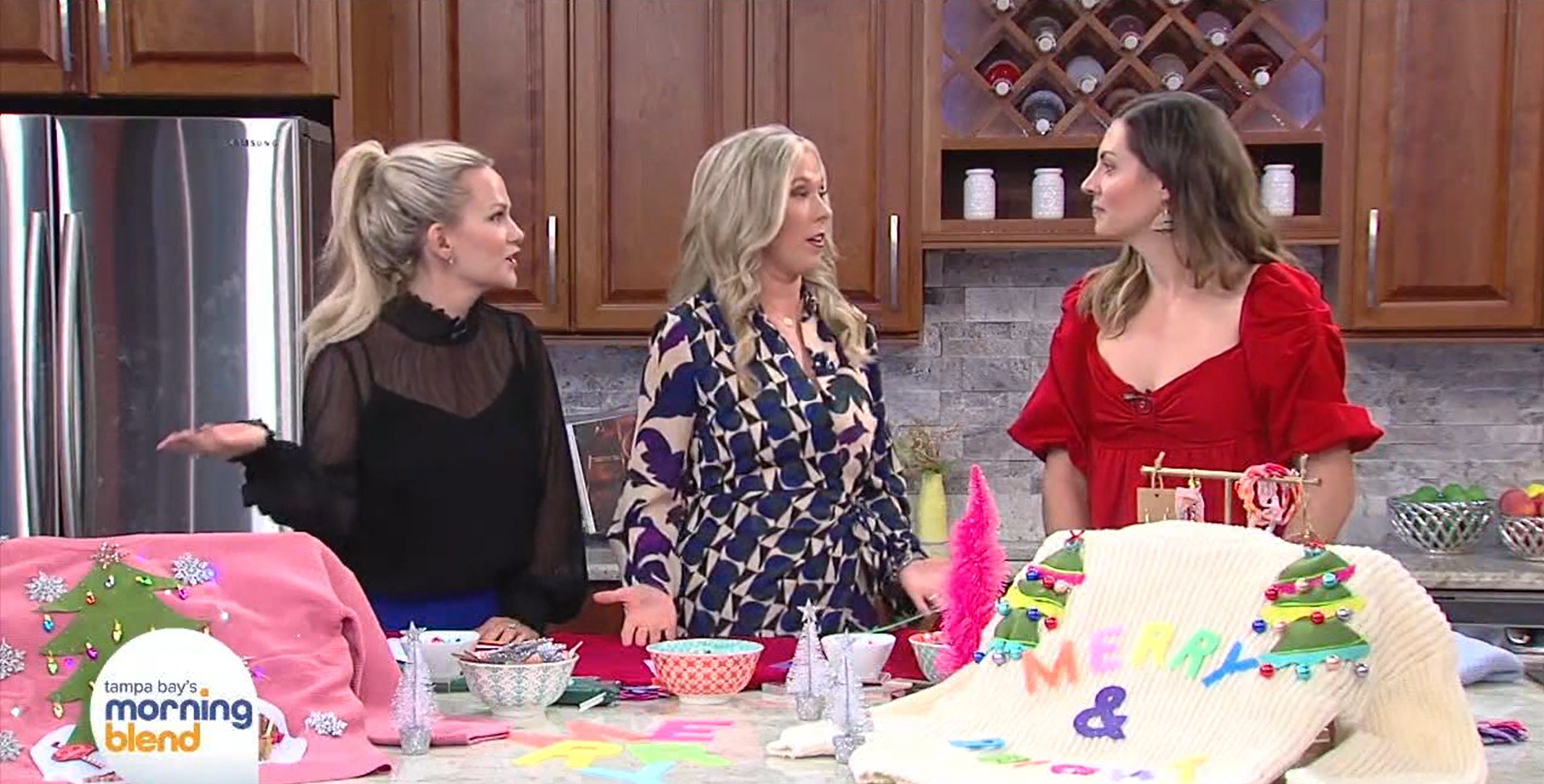 Three women are standing in a kitchen with wooden cabinets and a rustic backsplash, engaged in conversation. They are surrounded by colorful craft supplies—about sweaters, felt letters, and decorations—possibly for making holiday-themed crafts.