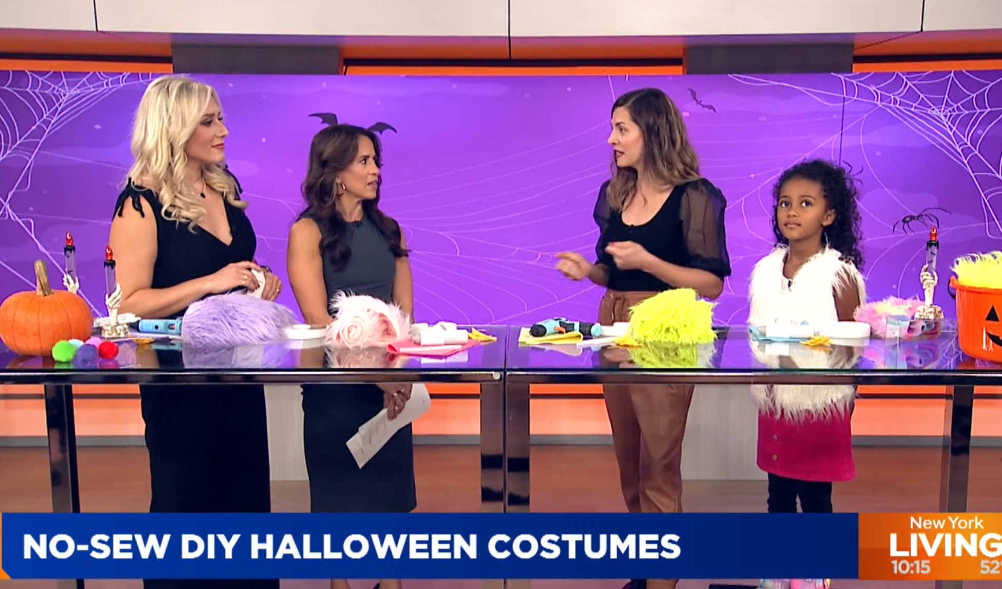 A group of four people, including a child, are standing behind a table with Halloween craft supplies. A spider web decoration is visible in the background. The text at the bottom reads, "NO-SEW DIY HALLOWEEN COSTUMES" and the left corner shows a timestamp "10:15" and "LIVE". This segment is all about creative costume making for Halloween.