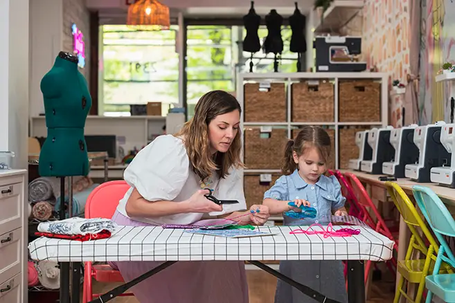 A woman and a young girl are working together on a craft project at a table in a brightly lit, colorful workshop that feels like home. The woman holds scissors while the girl is focused on the materials. The table is covered with fabric and sewing supplies, and there are sewing machines in the background.