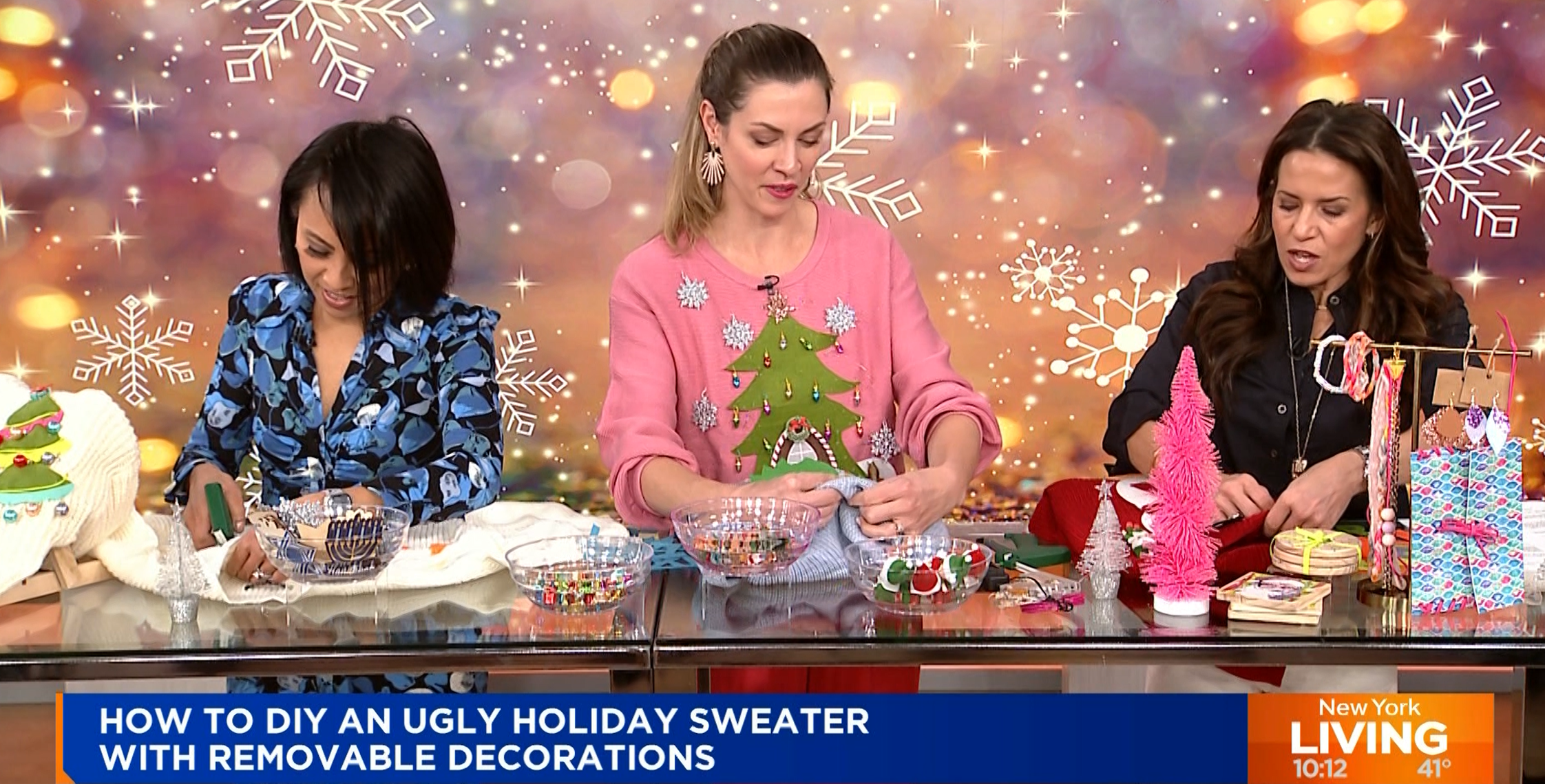 Three women sit at a table on a talk show, crafting Christmas sweaters with various decorations. The background features festive lights and snowflakes. The screen displays the text, "HOW TO DIY AN UGLY HOLIDAY SWEATER WITH REMOVABLE DECORATIONS." It’s all about getting creative this season!