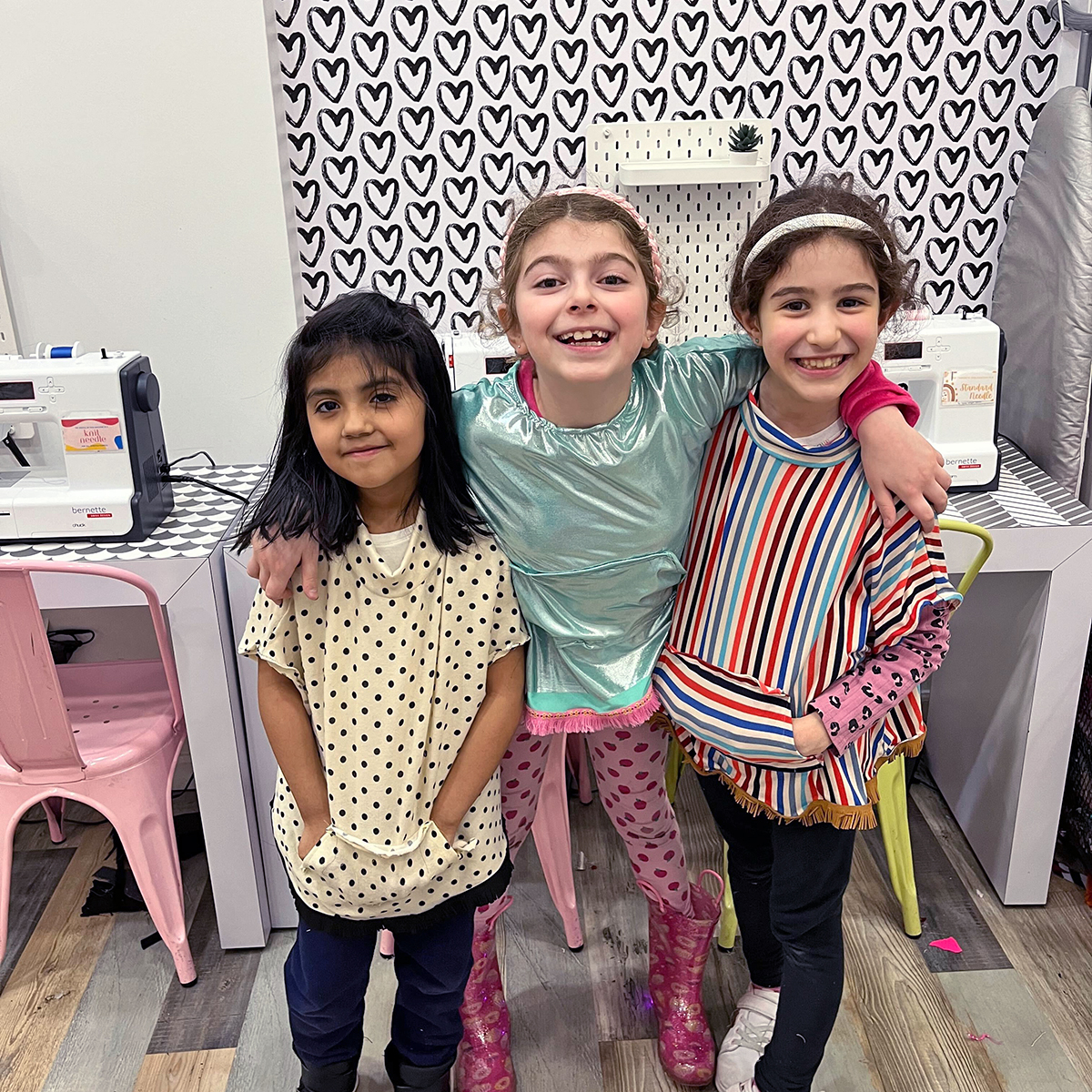 Three young girls stand together smiling in front of sewing machines. The girl on the left wears a polka dot shirt, the girl in the middle wears a green top and pink pants, and the girl on the right wears a colorful striped shirt. The background is decorated with heart patterns—steps to ownership for their creativity blossoming.