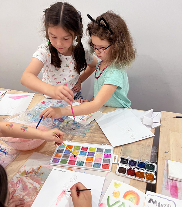 Two young girls are focused on painting with watercolors at a table. One girl in a white shirt with floral patterns dips her brush into paint, while the other girl in a light green shirt and cat ears reaches over. Scattered around are papers, brushes, and paint palettes—their steps to ownership of creativity clear.