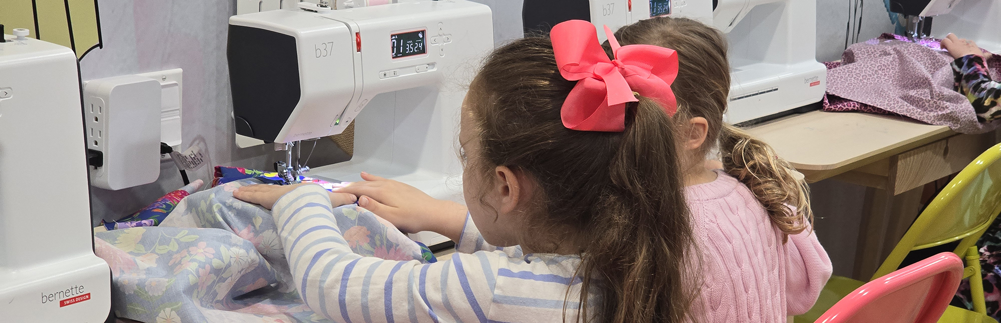 Two young girls sit side-by-side in front of sewing machines, concentrating on their sewing projects. One girl, with a large pink bow in her hair, is about to finish her piece. They are both working with colorful fabric pieces under the machine needles.