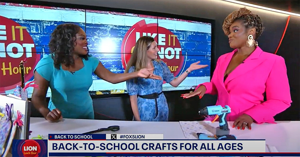 Three women are engaged in a lively conversation on a TV show set decorated with a colorful backdrop. A banner at the bottom of the screen reads: "BACK-TO-SCHOOL CRAFTS FOR ALL AGES." Craft supplies, including a glue gun, are visible in the foreground, offering viewers useful website information for their projects.