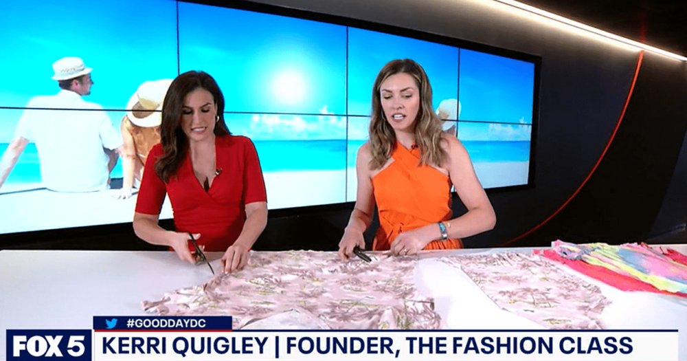 Two women are featured on a TV show set with an ocean scene backdrop. They are cutting fabric on a table. A banner at the bottom reads "FOX 5," "#GOODDAYDC," "Kerri Quigley | Founder, The Fashion Class." About the scene, one woman wears a red dress; the other, an orange dress.